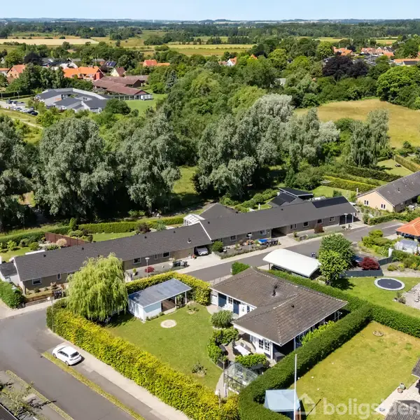 Rækkehuse med sorte tage står i en række. De omgivende haver er velplejede. Landskabet strækker sig op til trælunde og marker i baggrunden.