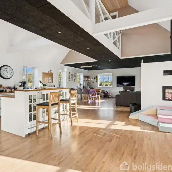 Et moderne åbent køkkenalrum med hvidt interiør, højloftede trælofter og trægulv. Der er en hvid pejs, barstole ved køkkenøen og et stort TV i stuen.