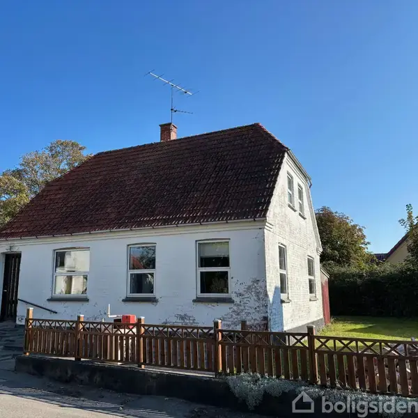 Hvidt hus med rødt tag står stille bag et træhegn i en have. Sollys oplyser facaden, mens en blå himmel danner baggrunden. Antenne på taget.