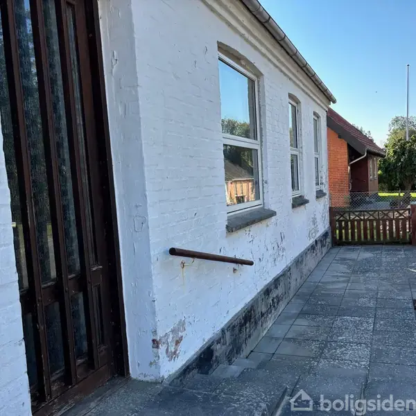 Hvidt murstenshus med vinduer og en dør i træ. Det står på en flisebelagt terrasse ved siden af en lille have med buske og en rød bygning i baggrunden.