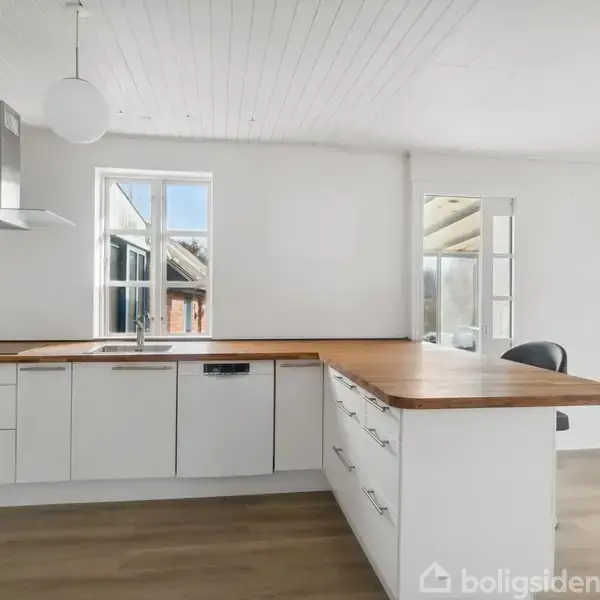 Et lyst køkken med hvide skabe og træbordplade. Køkkenet har to vinduer og en emhætte over komfuret. Trægulv og hvidt loft med en rund lampe.