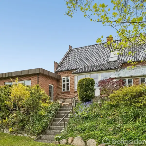 Et rødt murstenshus med sort tag står omgivet af en grøn have med buske og blomster. En trappe fører op til hoveddøren, mens himlen er blå i baggrunden.