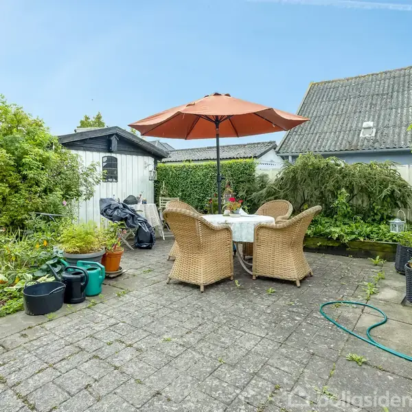 Et rundt bord med fire flettede stole står under en orange parasol på en flisebelagt terrasse. Omgivet af grønne planter, blomsterkrukker og en haveslange.
