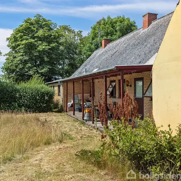 Et ældre stenhus med stråtag står i en have med højt græs og buske. Huset har en veranda med forskellige genstande og ligger omgivet af grønne træer under en blå himmel.