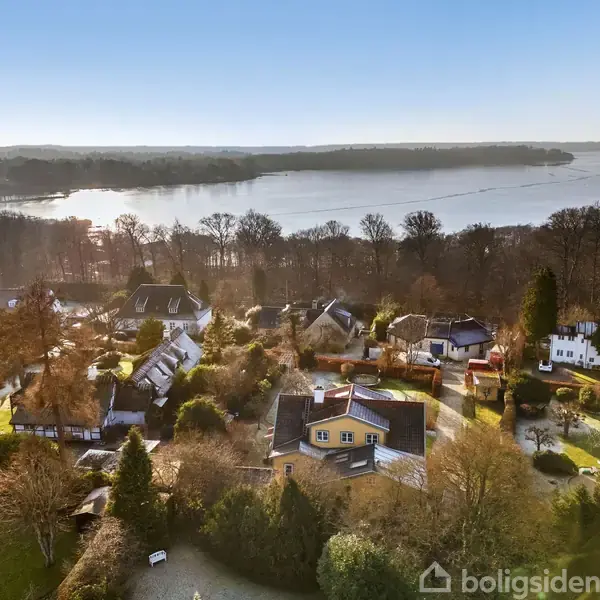 En række huse ligger stille blandt træer og haver ved siden af en sø, der spejler himmelen. Vandet strækker sig mod horisonten, omgivet af skovklædte områder.