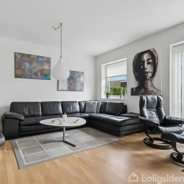 Sort lædersofa står i en stue med trægulv, omgivet af moderne kunst på væggene. Store vinduer lukker lys ind, og en sort lænestol står ved siden af et sofabord.