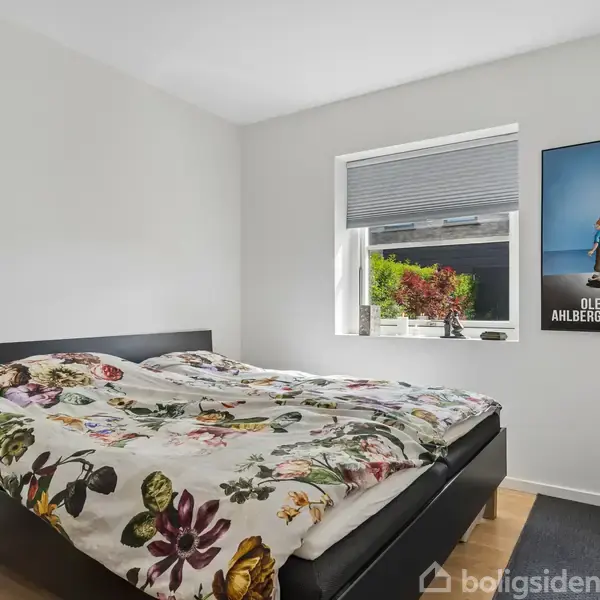 Sengen er dækket med et blomster mønstret sengetæppe, står i et rum med lyse vægge. Ved siden af et vindue hænger en plakat med teksten "OLE AHLBERG".