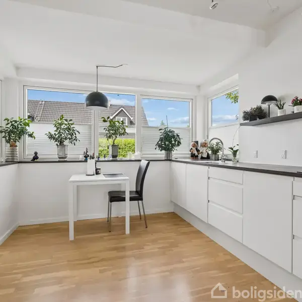 Lyst, moderne køkken med hvide skabe og mørke bordplader. Enkelt bord med stol mod store vinduer, der giver udsigt til træer og nabohus. Grønne planter pynter i rummet.