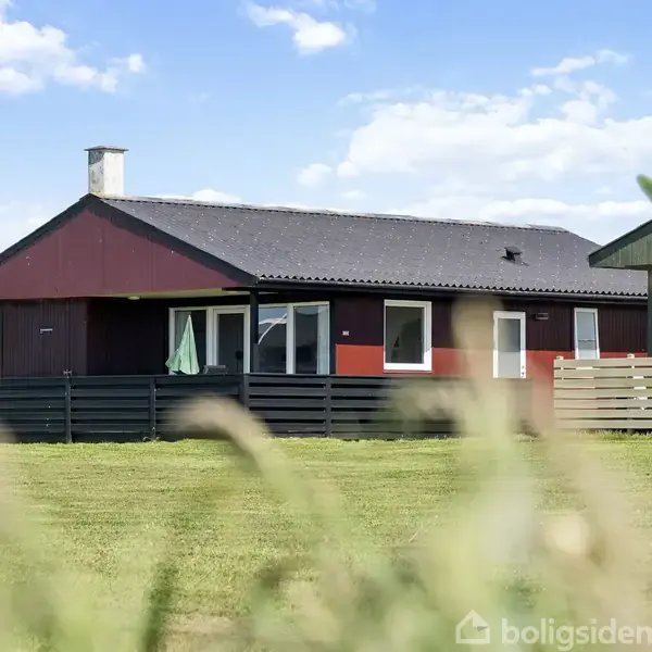 Et rødt hus står stille mellem andre bygninger i et grønt område med en blå himmel i baggrunden. En sort træterrasse omringer huset.