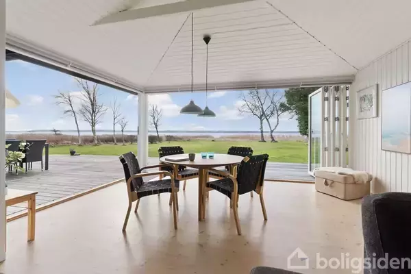 Et rundt spisebord med stole står på et lyst trægulv inde i et rum med åbne døre ud mod en terrasse. Udenfor ses en stor græsplæne og havudsigt.