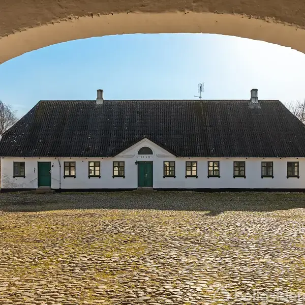 En hvid landbygning med mørkt tag står centralt, omgivet af en brostensbelagt gårdsplads. Bygningen ses gennem en buet portåbning. Himmel og træer i baggrunden.