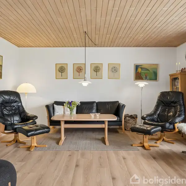 Stue med sorte læderlænestole og sofa, der står omkring et træbord. Der er lamper, billeder på væggen og et stort vindue med gardiner, der skaber et lyst rum.