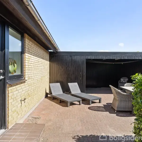 Terrasse med to liggestole og fletmøbler står på en flisebelagt gårdhave foran et murstenshus. I baggrunden ses en sort træbygning samt grønne buske under en blå himmel.