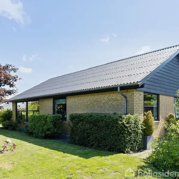 Et gult murstenshus med skråt tag står i en velplejet have omgivet af grønne buske og træer under en klar blå himmel.