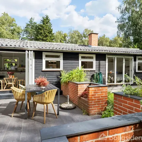 Sort, træbeklædt hus med store vinduer, hvidrammede terrassedøre og en stenbelagt terrasse med træmøbler. Omgivet af en frodig, grøn have med høje træer i baggrunden.