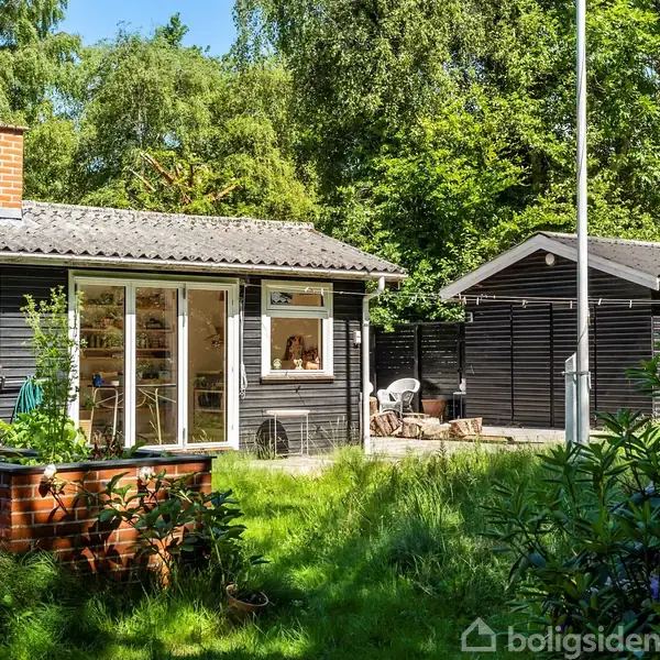 Sort træhus med store vinduer står ved en terrasse med bord og stole, omgivet af grønne buske og træer i en rummelig have.