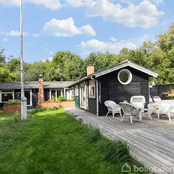 Sort træhus med runde vinduer står på en træterrasse med hvide møbler. Græsplæne omgiver huset; omgivet af tæt, grøn skov under en blå himmel med skyer.