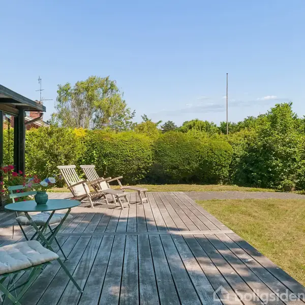 Rundt spisebord med sorte stole står i et træbeklædt rum. En loftlampe hænger over bordet. Store vinduer og en glasdør viser en terrasse udenfor.