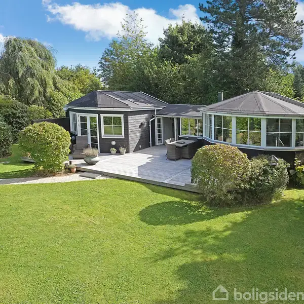 Et sortmalet hus med en stor terrasse omgivet af grønne buske og træer. Det ligger i en frodig have under en blå himmel med lette skyer.