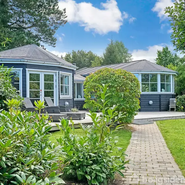 Et moderne, sortmalet hus med store vinduer står stille omkranset af frodige grønne træer og buske. En belagt sti fører gennem en velplejet græsplæne, under en blå himmel.