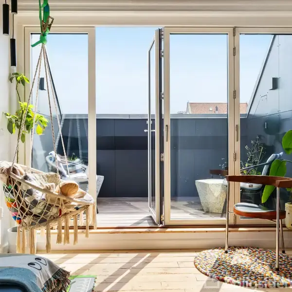 Hængestol svæver ved siden af åbne altandøre i et lyst rum med planter, guitar og drejestol. Døre fører til en lille altan i baggrunden.