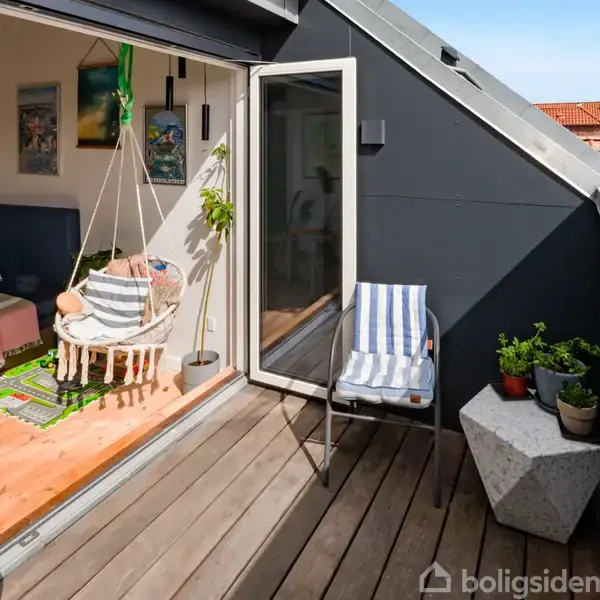En altan med en stribet stol og planter ved siden af et moderne loftværelse. Værelset har gyngestol og sofa, adskilt af en glasskydedør. Solens lys fylder rummet.