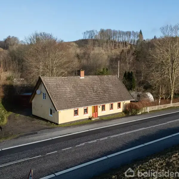 Et gult hus med saddeltag står ved en vej, omgivet af træer og bakker i vinterlandskab. En bil er parkeret til venstre ved siden af huset.