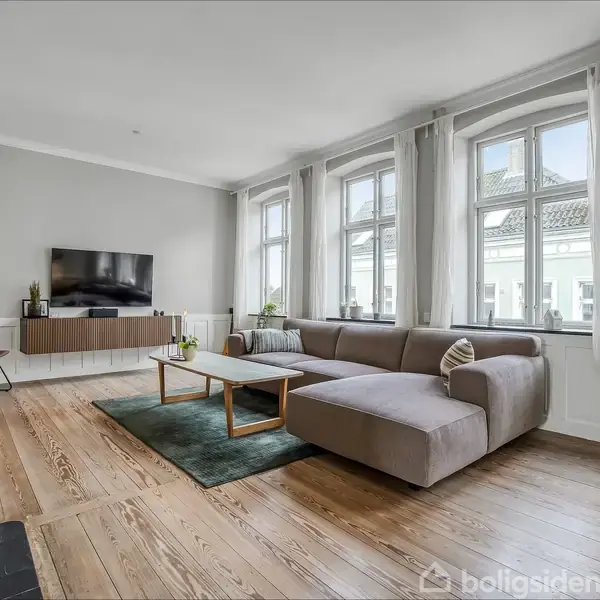 Et moderne opholdsrum med en stor, grå hjørnesofa ved siden af fem vinduer. Et fladskærms-tv hænger på væggen, og et sofabord står på et tæppe.