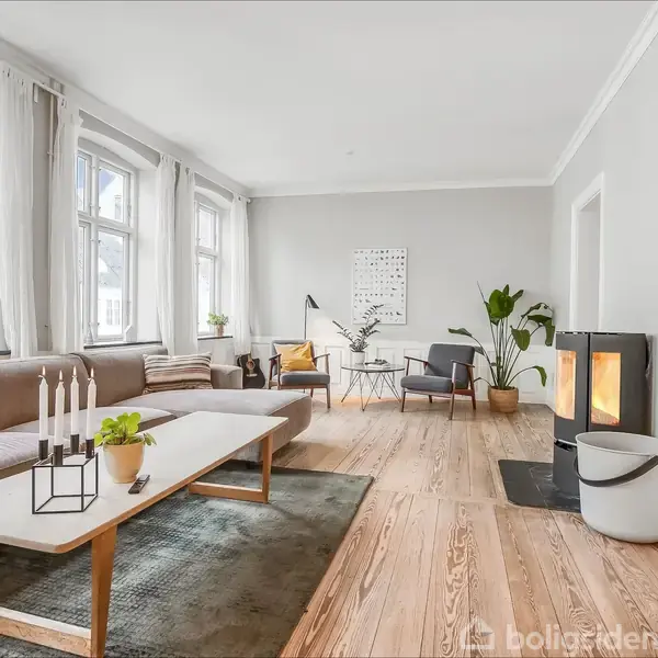 En lys stue med en grå sofa ved et stort vindue, et hvidt bord med en plante, to stole omkring et bord, og en brændeovn ved siden af en plante.