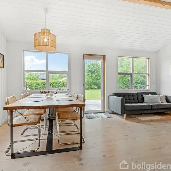 Et spisebord med dækkede tallerkener står ved siden af en sofa i en lys stue. Store vinduer og en glasdør leder ud til en grøn have.