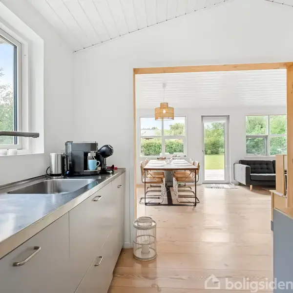 Køkkenvask med kaffemaskine står stille i lyst køkken, åbnes mod spisestue med bord og stole, omgivet af store vinduer, der viser grøn have udenfor.