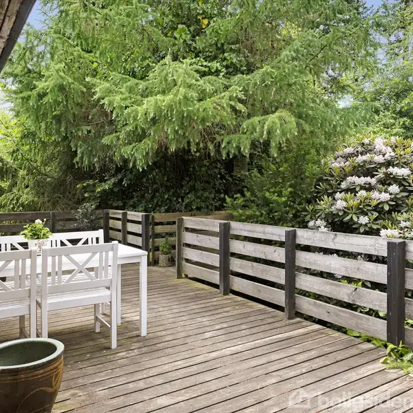 Et hvidt bord og stole står på en træterrasse. Terrassen er omgivet af grønne planter og høje træer, der skaber en naturlig og privat atmosfære.