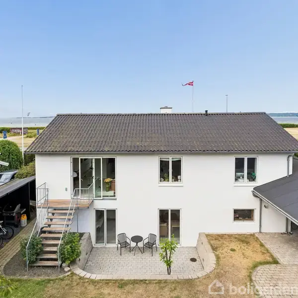 Et hvidt to-etagers hus med fladt, gråt tag står stille. Huset omgives af en lille gårdhave med bord og stole, og i baggrunden ses kysten og havet.