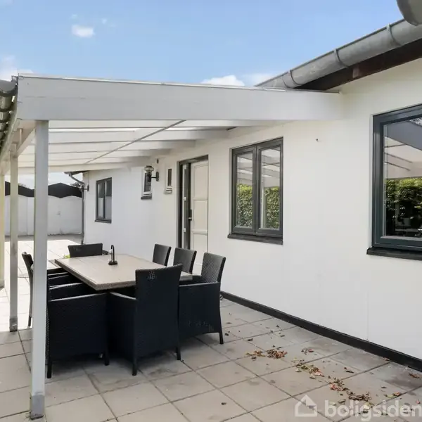 Terrasse med sort spisebord og seks stole under et halvtag. Omgivet af en hvid murstensvæg og flisebelægning, med grønne hække til venstre.