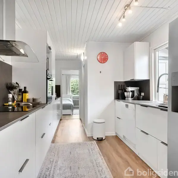 Et moderne køkken med hvide skabe og stålapparater ligger i en lys gang. Der er en rød vægur på væggen og en åbning til en stue med sofa.