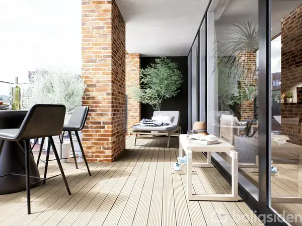 Et terrasseområde med træplanker, hvor to stole og et bord står nær murstensvægge. Planter og en liggestol ses længere væk ved store glasvinduer. En afslappet udendørs atmosfære.