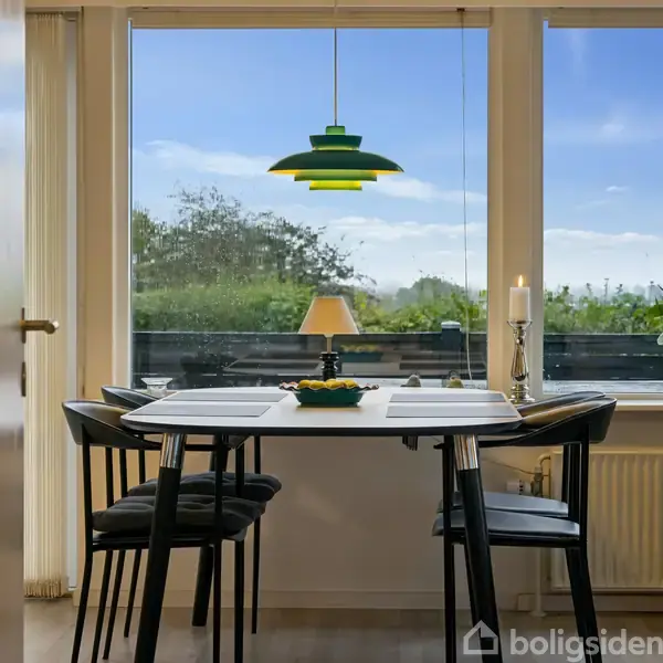 Et spisebord med fire stole står i et lyst rum. En grøn pendellampe hænger over bordet. Gennem store vinduer ses en have under en blå himmel.