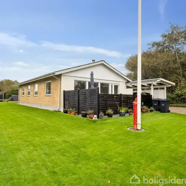 Et gult murstenshus står stille omgivet af en velholdt grøn græsplæne og små træer. Der ses en overdækket terrasse og en grusbelagt indkørsel i baggrunden.