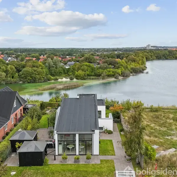 Moderne hus med fladt tag og store vinduer ligger ved en sø. Græsplæne og terrasser omgiver huset. I baggrunden ses skov, flere huse og en bysilhuet.