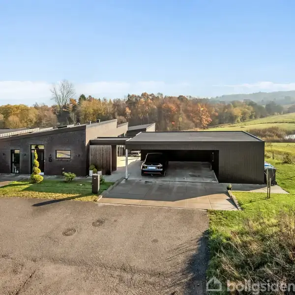 En moderne bolig med fladt tag og carport, hvor en bil holder parkeret, placeret i et naturskønt område med grønne træer og en åben mark i baggrunden.