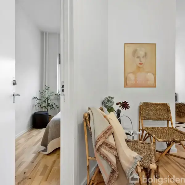 To flettede stole med tæpper placeret i en lys gang. En billedramme hænger på væggen, og i baggrunden ses en plante i et rum med trægulve.