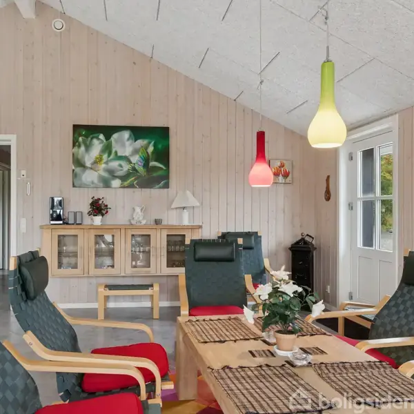 Stue med to grønne lænestole omkring et sofabord, dekoreret med blomster. Vægge i træpaneler, en reol med nips, og farverige lamper hængende. Dør fører til udendørs område.
