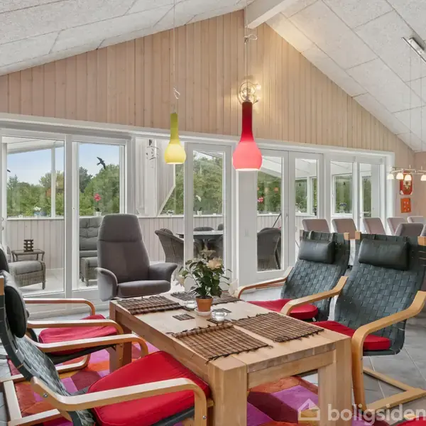 Stue med spisebord og to sofagrupper, belyst af farverige lamper. Store vinduer giver udsigt til en terrasse og skovomgivelser. Møblerne er komfortabelt opstillet til afslapning og samvær.