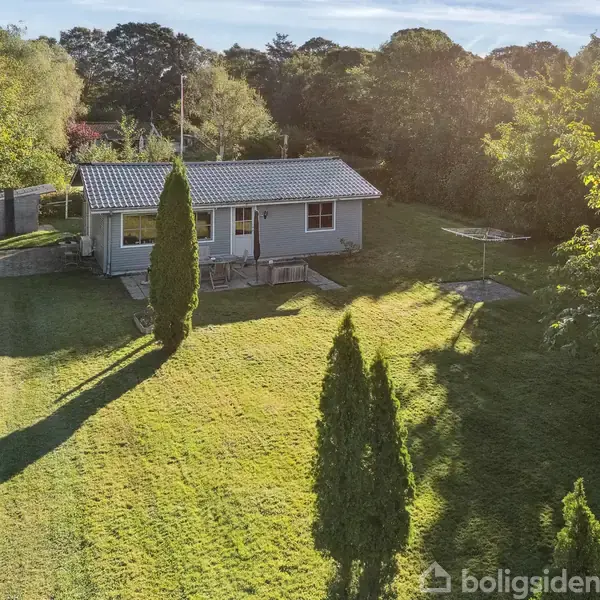 Lille gråt hus med fladt tag står i solrig have omgivet af høje træer. En flagstang er placeret foran huset, som er omgivet af grønne buske og skygger på græsplænen.