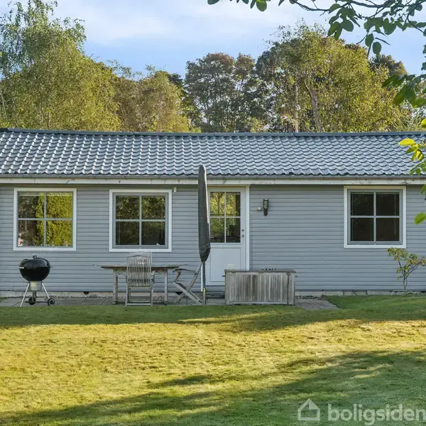 Et gråt sommerhus med tre vinduer og en dør står i en have. En grill og havemøbler er foran huset, omgivet af græs og træer i baggrunden.