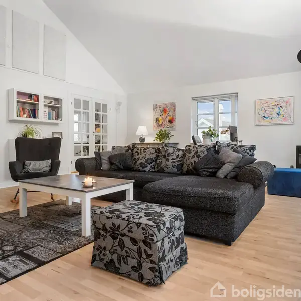 En stor, grå sofa med mønstrede puder står i en lys stue. En sort stol og sofabord er til venstre, ved siden af et brændekomfur og vinduer. To malerier pryder væggene.