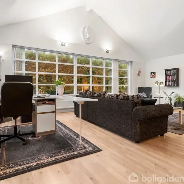 Et lyst stuerum med trægulv, sort sofaarrangement og skrivebord med kontorstol. Store vinduer fylder væggen, lukker naturen udenfor ind i rummet. Vægur hænger over sofaen, fjernsyn på væggen.