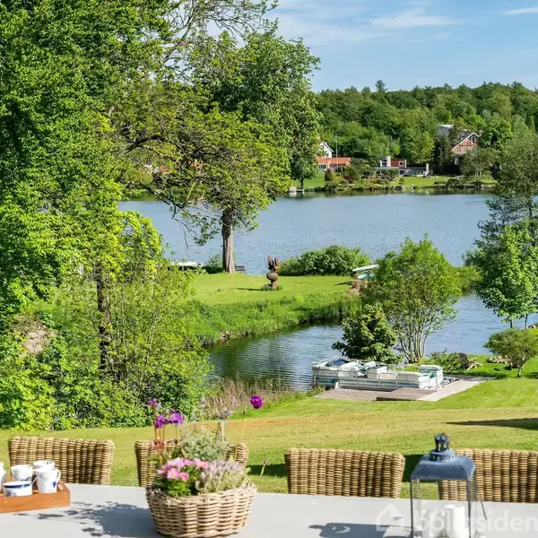 En terrasse med kurvestole og planter i forgrunden. Udsigt over en have, en flod med både og en skovklædt baggrund. Træer og huse ses i det fjerne.