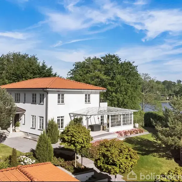 En stor hvid villa med rødt tegltag står stille i en velplejet have nær en sø. Omkring det grønne område findes buske, træer og en klar blå himmel.