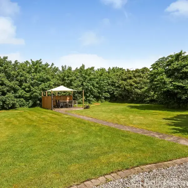 En pavillon står på en græsplæne, belagt med brosten, omkranset af frodige træer under en klar blå himmel. Ingen tekst er til stede.